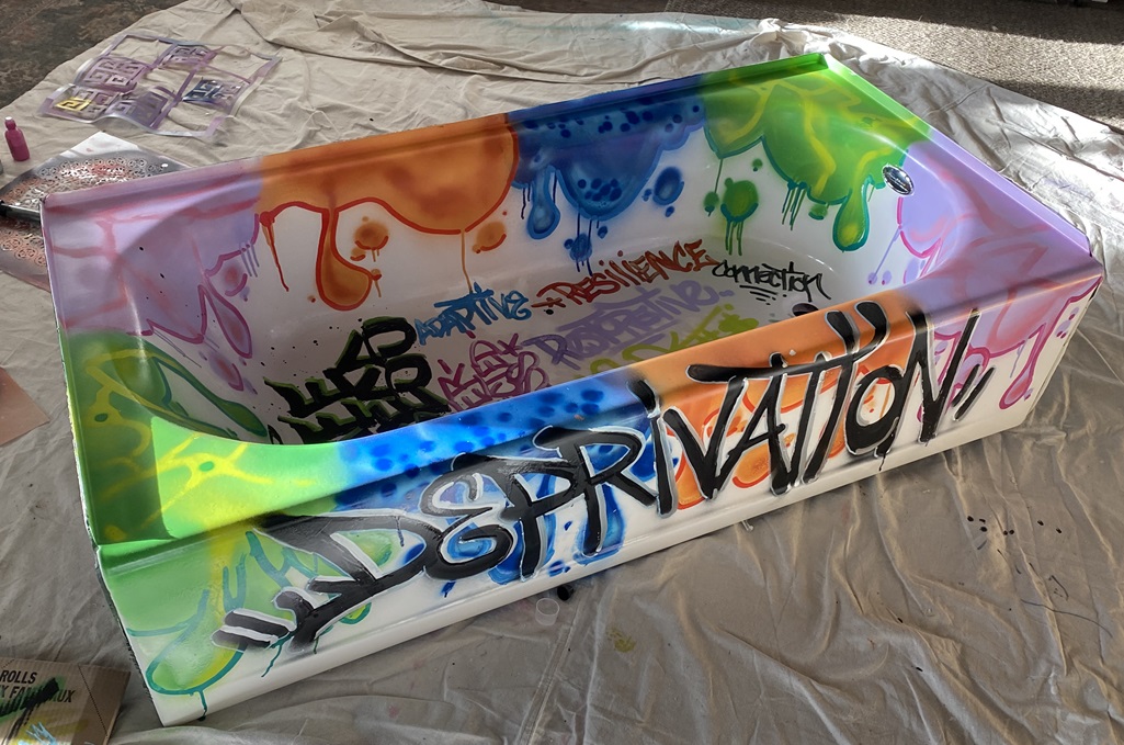 Brightly graffiti painted bathtub with the word "Deprivation" on its side and words like Resilience, Connection, and Restorative on the inside